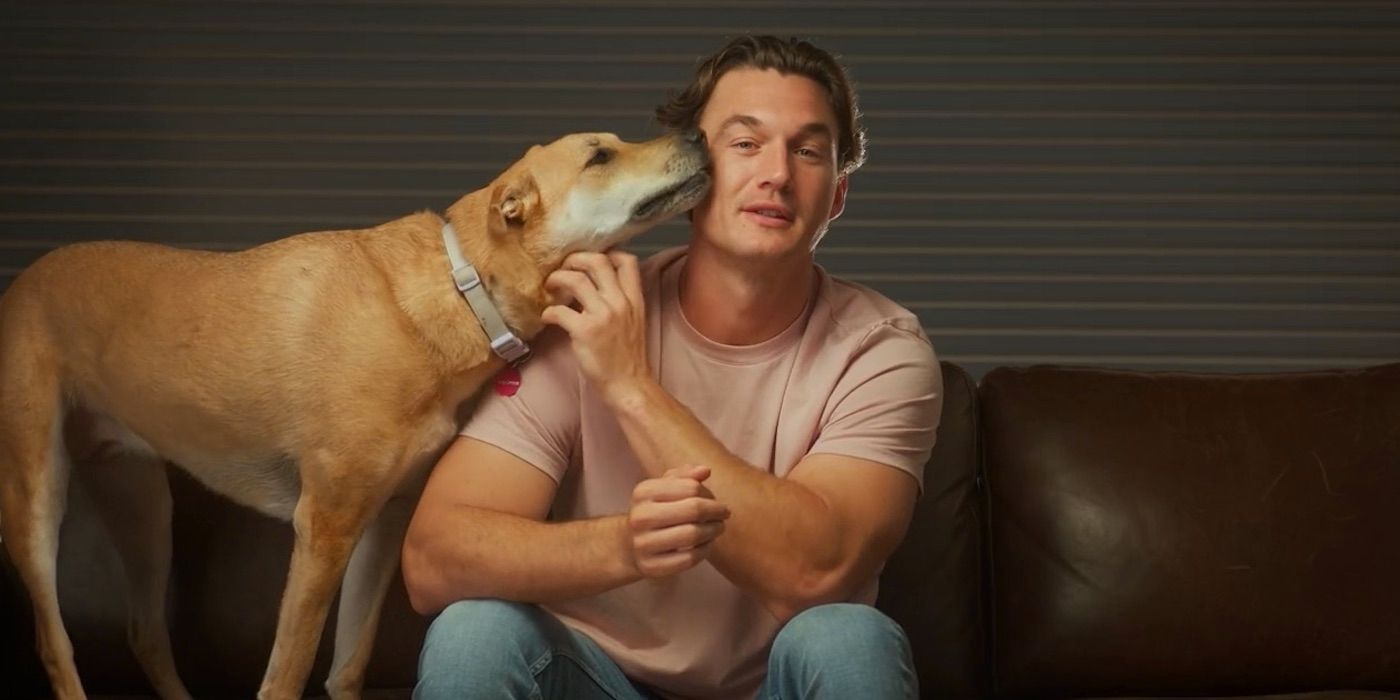 Tyler Cameron sits on a couch, scratching his dog&rsquo;s chin while she licks his face