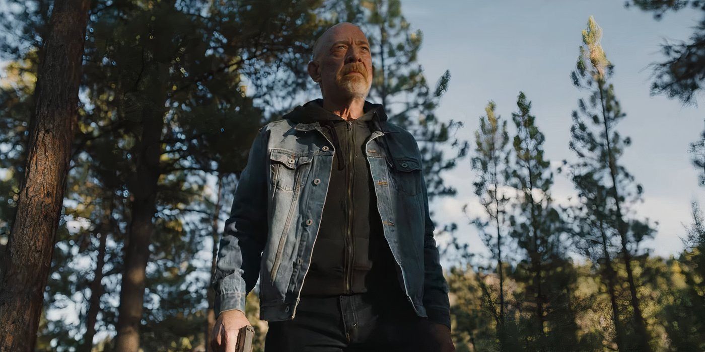 J.K. Simmons standing in the woods wearing a jean jacket clutching a pistol.