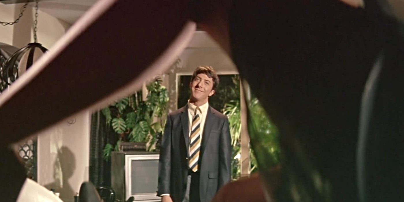 Dustin Hoffman as Benjamin Braddock, standing and smiling, framed by a woman's leg in The Graduate