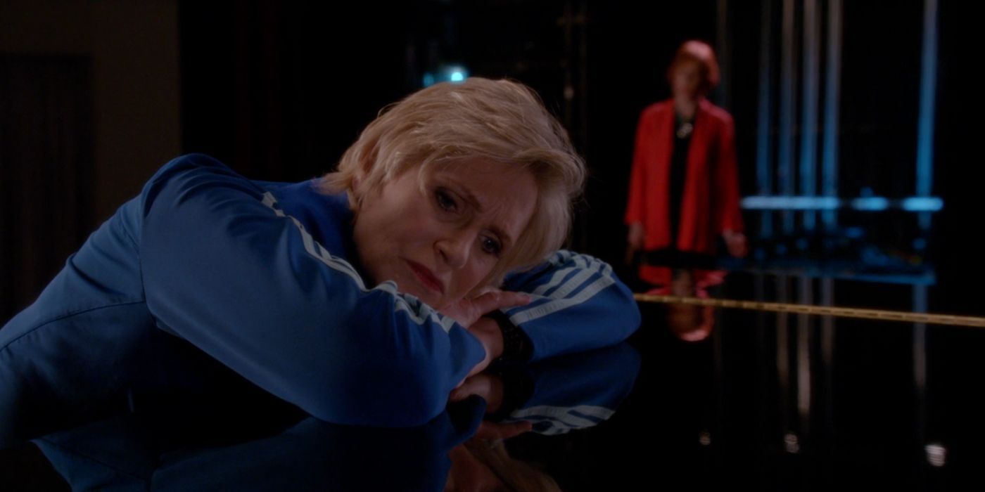 Sue Sylvester, wearing her signature blue track suit, rests her head on a black piano on a stage. Behind Sue, her red-headed mother, wearing a red coat and a black dress, is looking at her. 