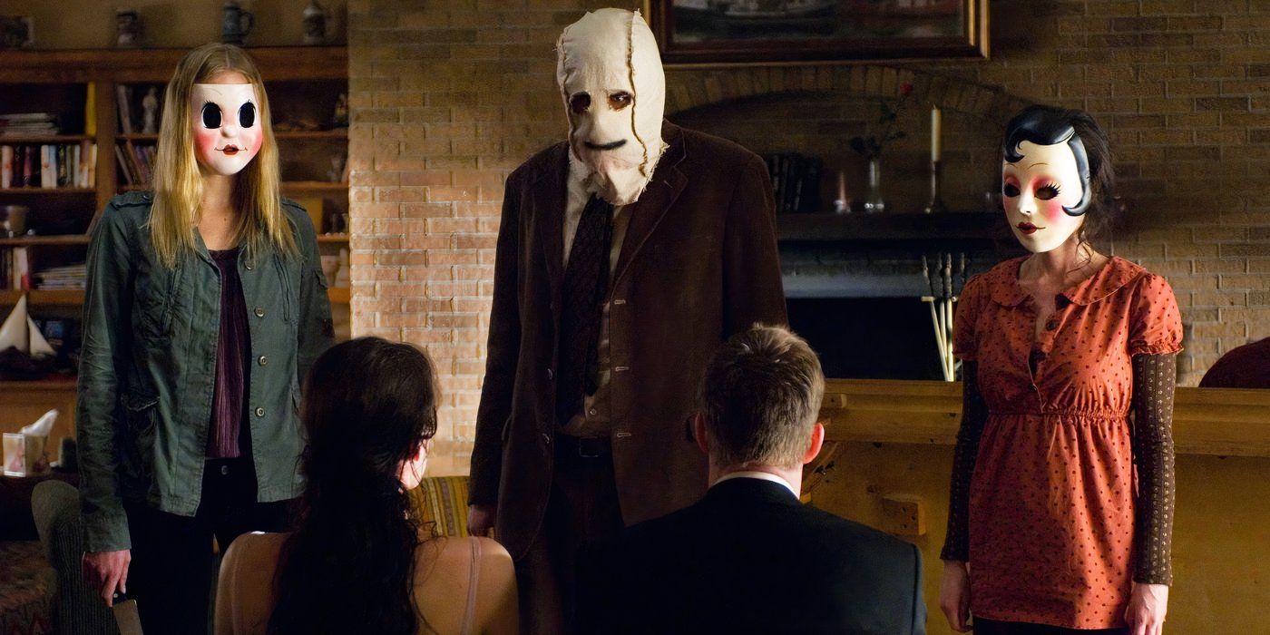 Pin-Up Girl (Laura Margolis), Man in the Mask (Kip Weeks), and Dollface (Gemma Ward) standing in a living room looking at Kristen (Liv Tyler) and James (Scott Speedman) who are tied to chairs in The Strangers.