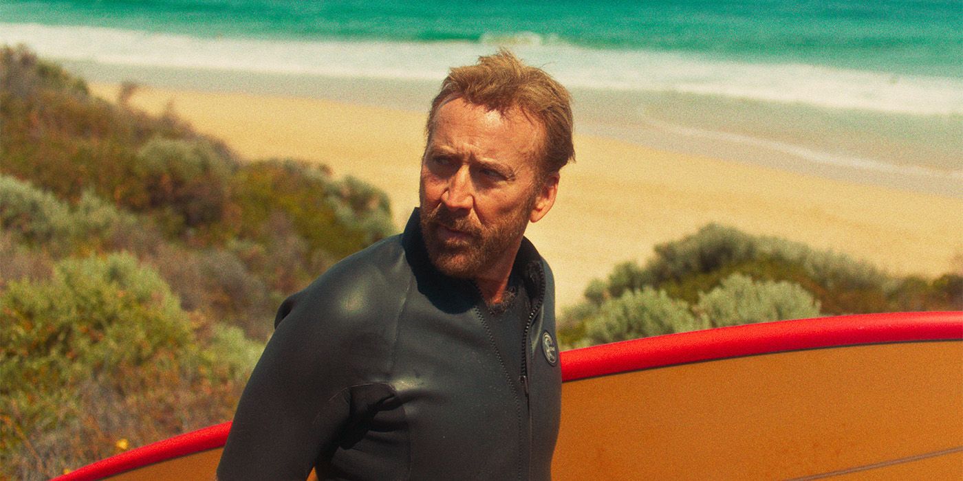 Nicolas Cage as The Surfer holding a surfboard on a beach looking over his shoulder