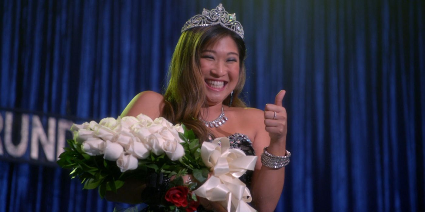 Tina smiles and gives a thumbs up to her friends offstage after she has been crowned prom queen. She is wearing a strapless sparkly black dress, silver jewelry, and a shiny silver crown. She is holding white roses.
