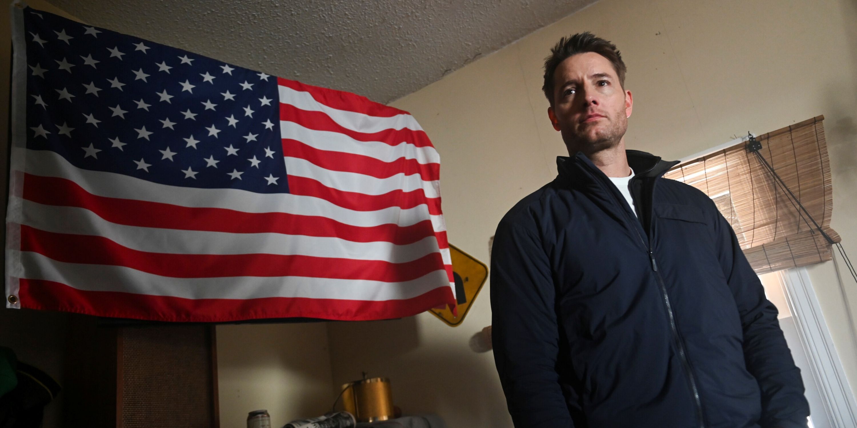 Justin Hartley as Colter Shaw standing in front of an American flag inside a house in the CBS series Tracker