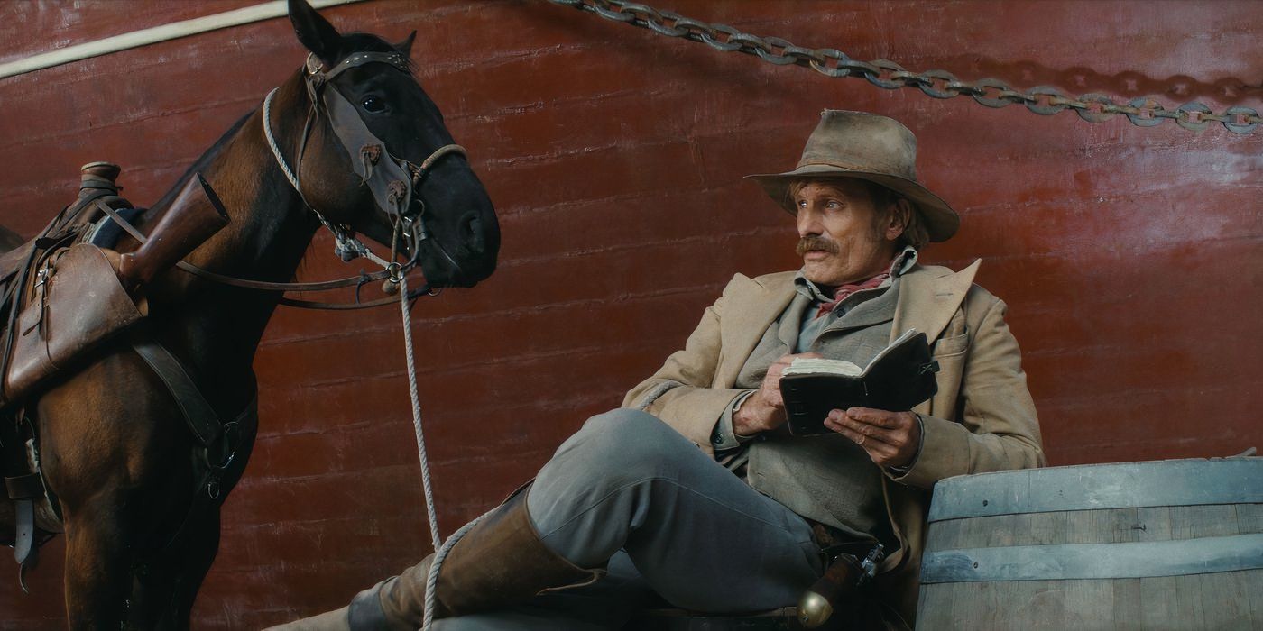 Viggo Mortesen in cowboy gear reading a book while sitting next to his brown horse.