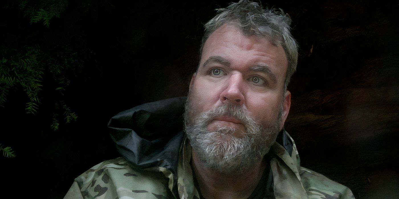 A bearded man wearing a camo jacket looks off camera, the dark woods behind him