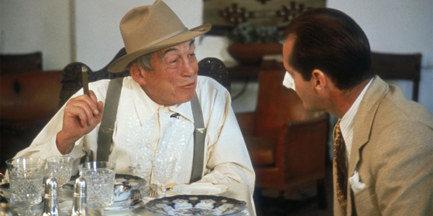 John Huston holding a cigar and talking amiably to Jack Nicholson at a table in a restaurant 