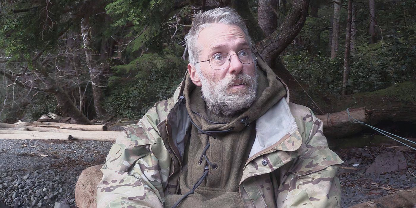 A man with glasses and a beard wearing camo outdoor gear sits on a rocky beach looking off camera