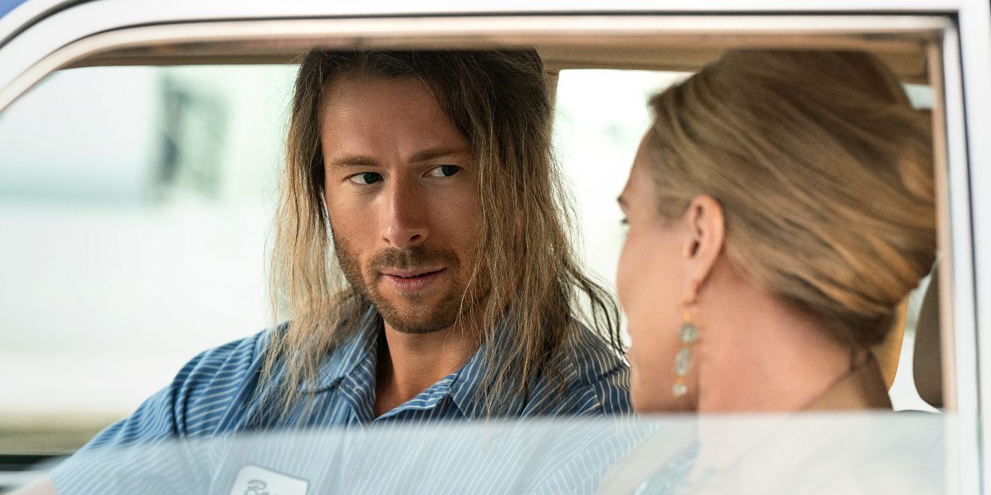 Glen Powell sitting in a passenger seat with long greasy hair.