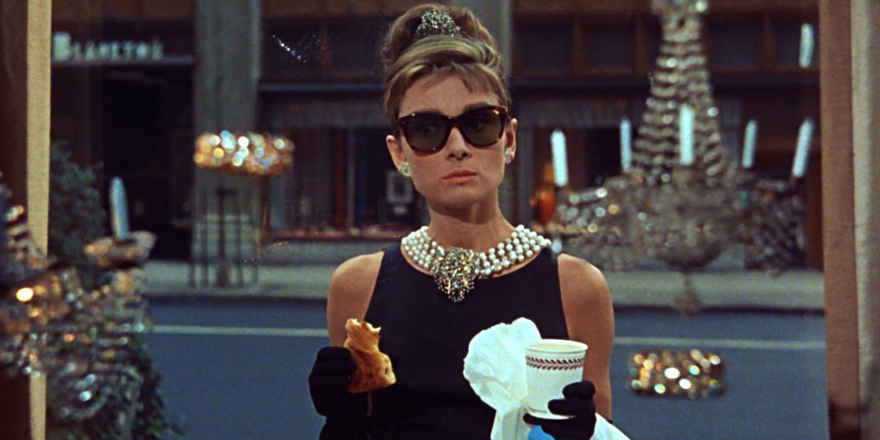 Holly in a black dress and tiara holding a coffee and a piece of pastry