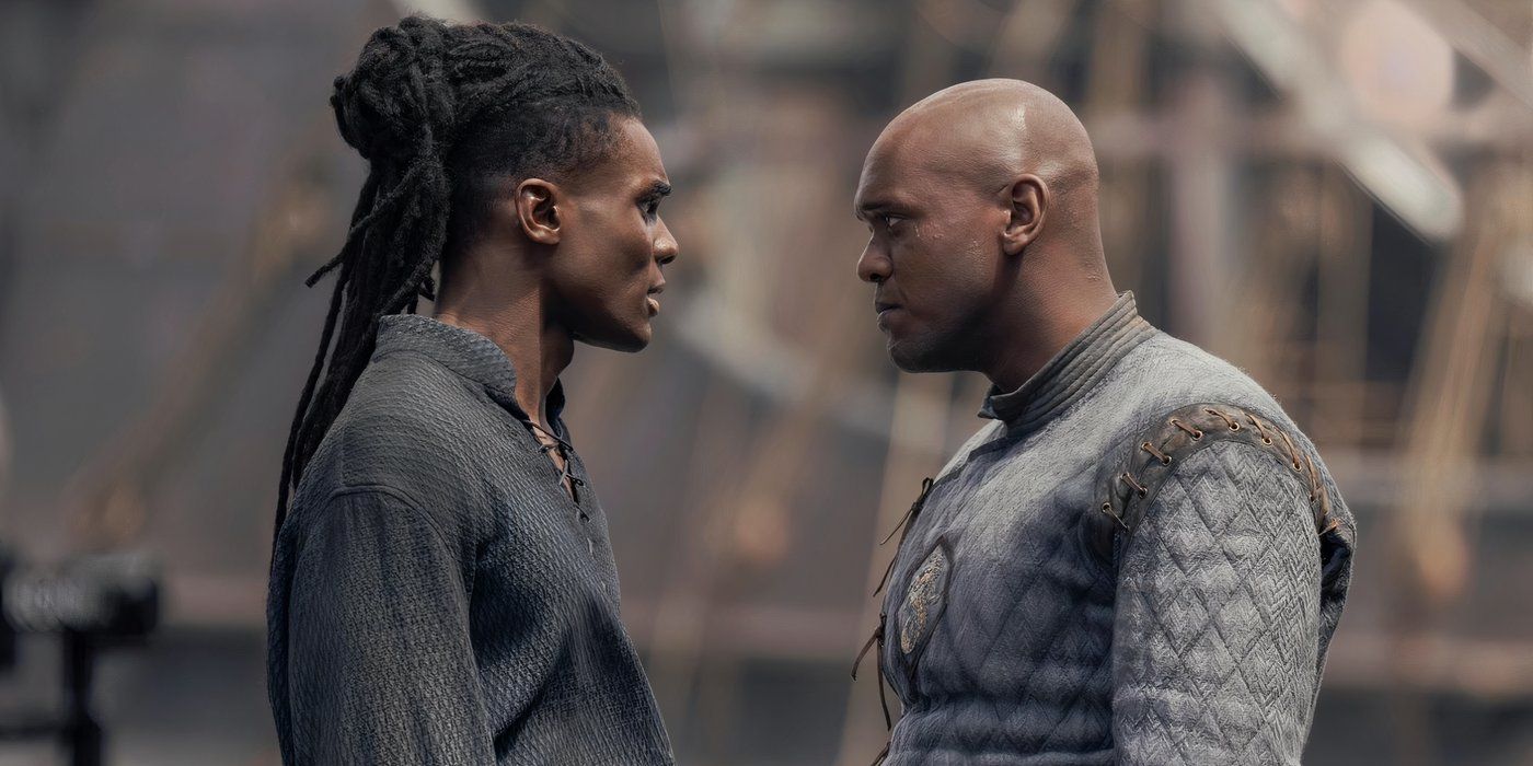 Two men face each other, one with a pile of black dreads and the other bald. They are both dressed in gray tunics and we can only see from their waist up.