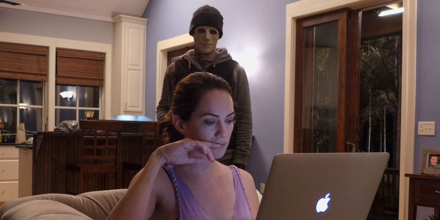 Maddie (Kate Siegel) sits at her laptop while a masked man (John Gallagher Jr.) stands behind her in Hush