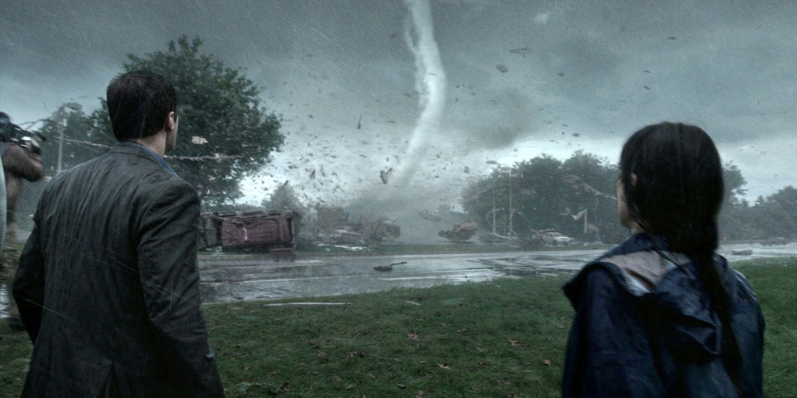 A man and a woman stand with their backs to the camera during a storm and watch a large tornado rip through the street infront of them