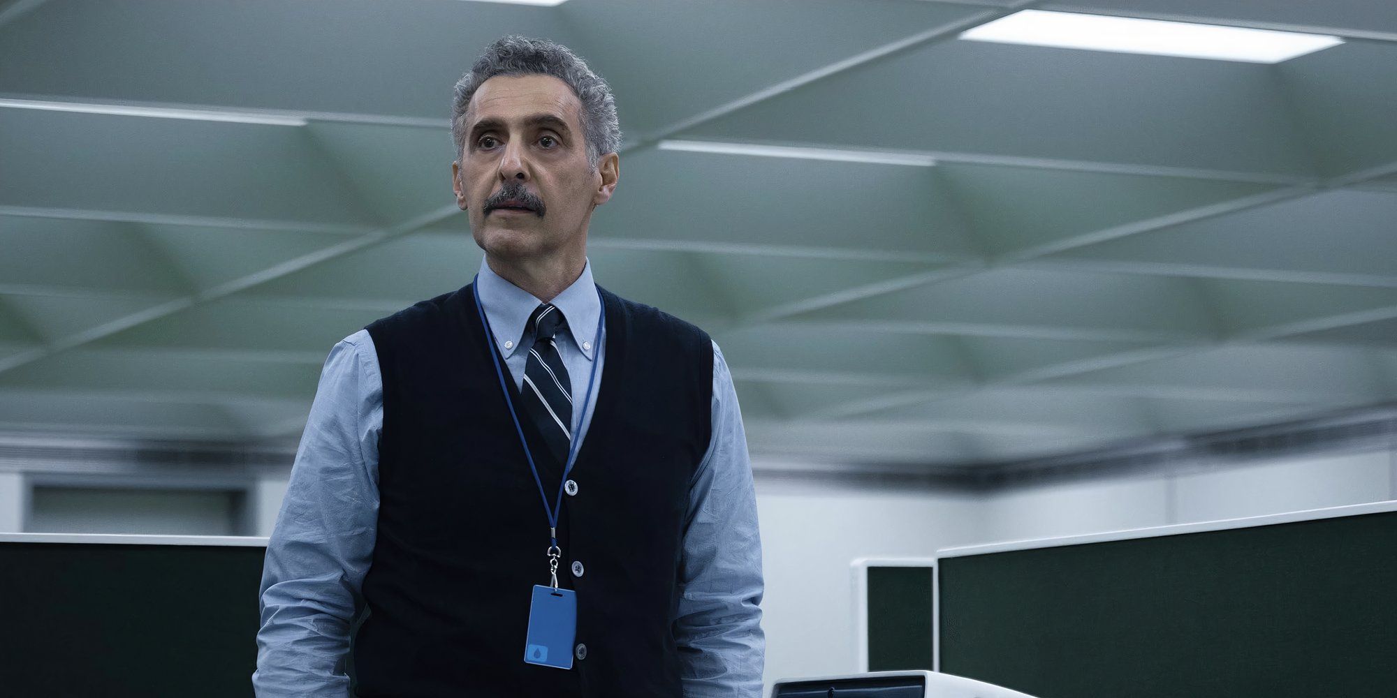 Irving B (John Turturro) stands in the Macrodata Refinement office wearing his work lanyard in Severance.