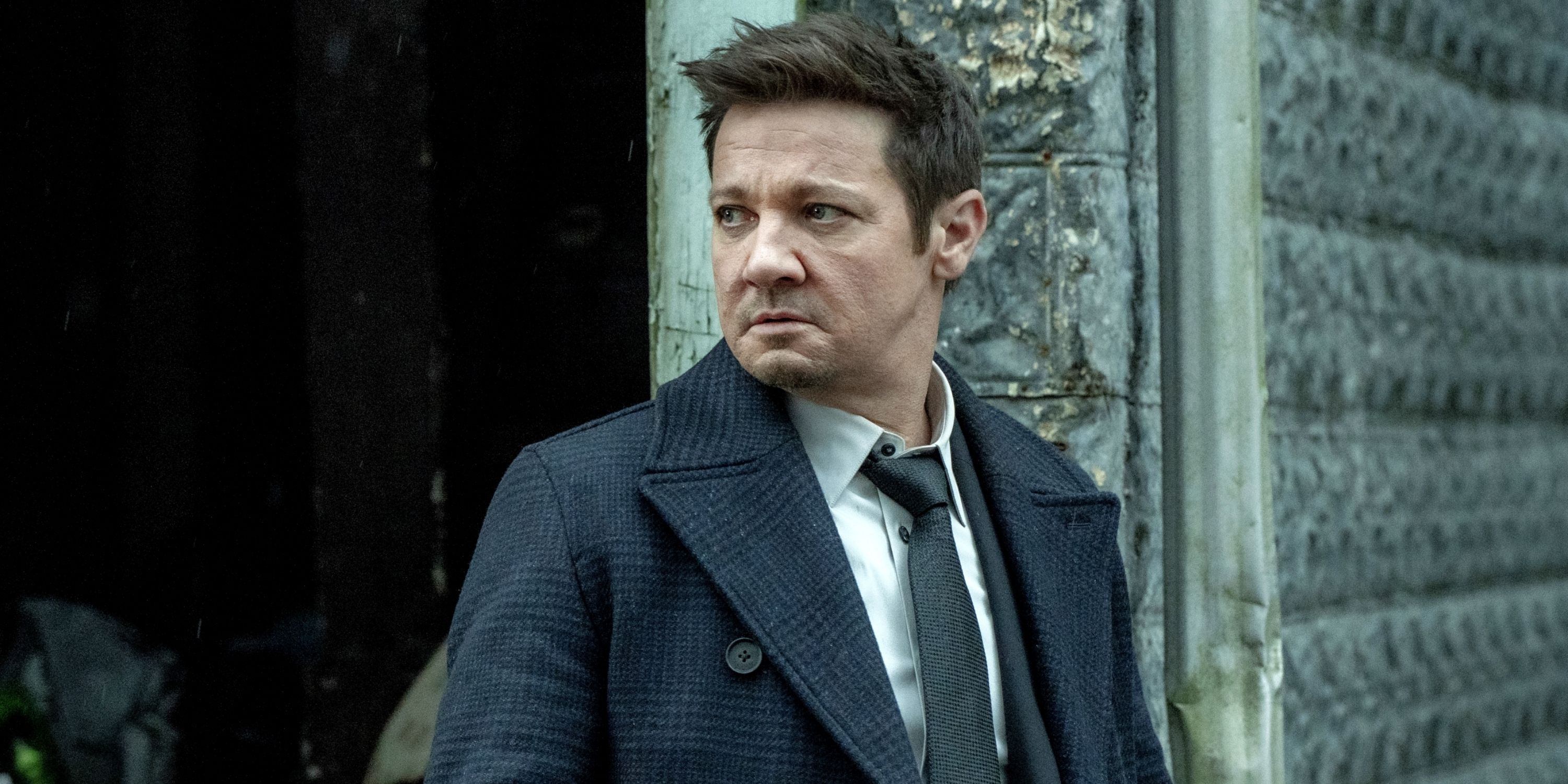 Jeremy Renner as Mike McLusky leaning against a wall in a suit and looking to the right in Mayor of Kingstown