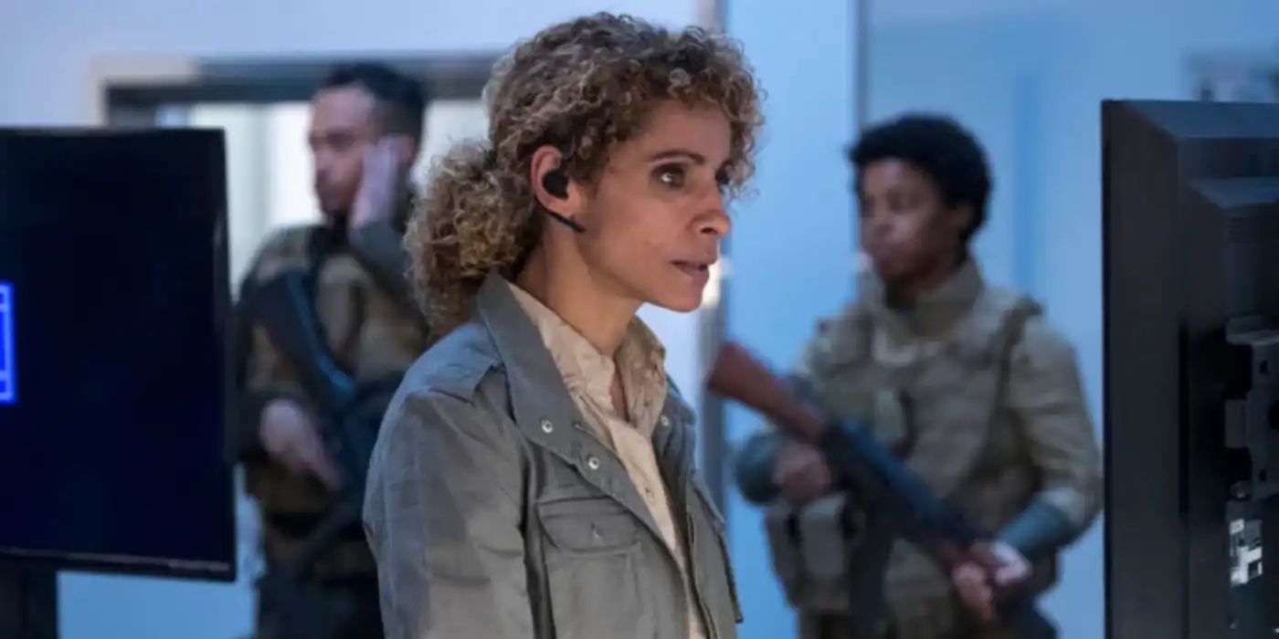 Shepherd types on a computer diligently as two armed guards stand behind her in military uniform.