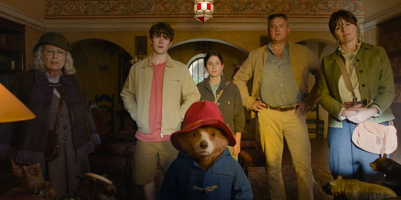 (left to right) Mrs. Bird (Julie Walters), Jonathan (Samuel Joslin), Paddington (Ben Wishaw), Judy (Madeleine Harris), Henry (Hugh Bonneville), and Mary (Sally Hawkins) standing in an office in 'Paddington in Peru'