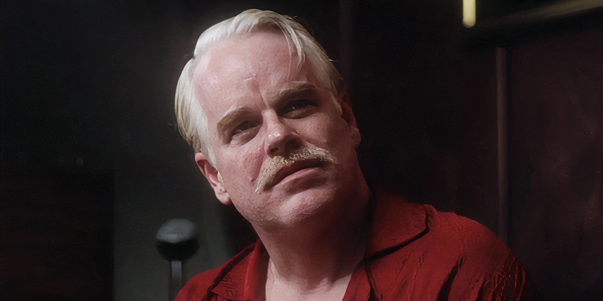 A closeup of Lancaster Dodd wearing a red shirt, a mustache, and an expression of deep thought