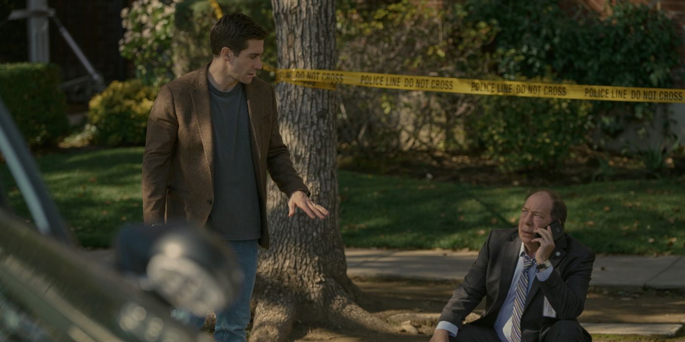 Jake Gyllenhaal está parado junto a un árbol con cinta adhesiva de advertencia al costado de la carretera, hablando con Bill Camp, quien está sentado y hablando por teléfono.