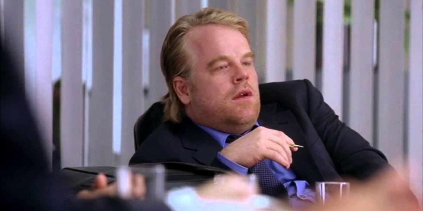 En Along Come Polly (204), Sandy Lyle (Philip Seymour Hoffman) se sienta incómoda en una silla de la sala de juntas con un traje.