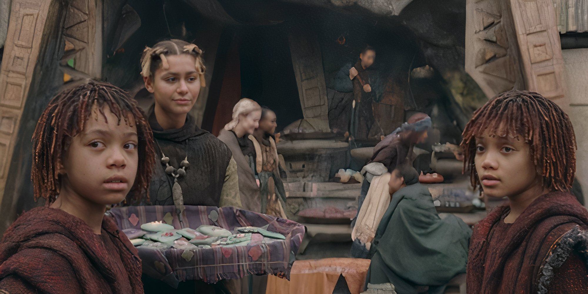 A young Osha and Mae look toward someone off screen while a woman holds a tray of treats in front of them.