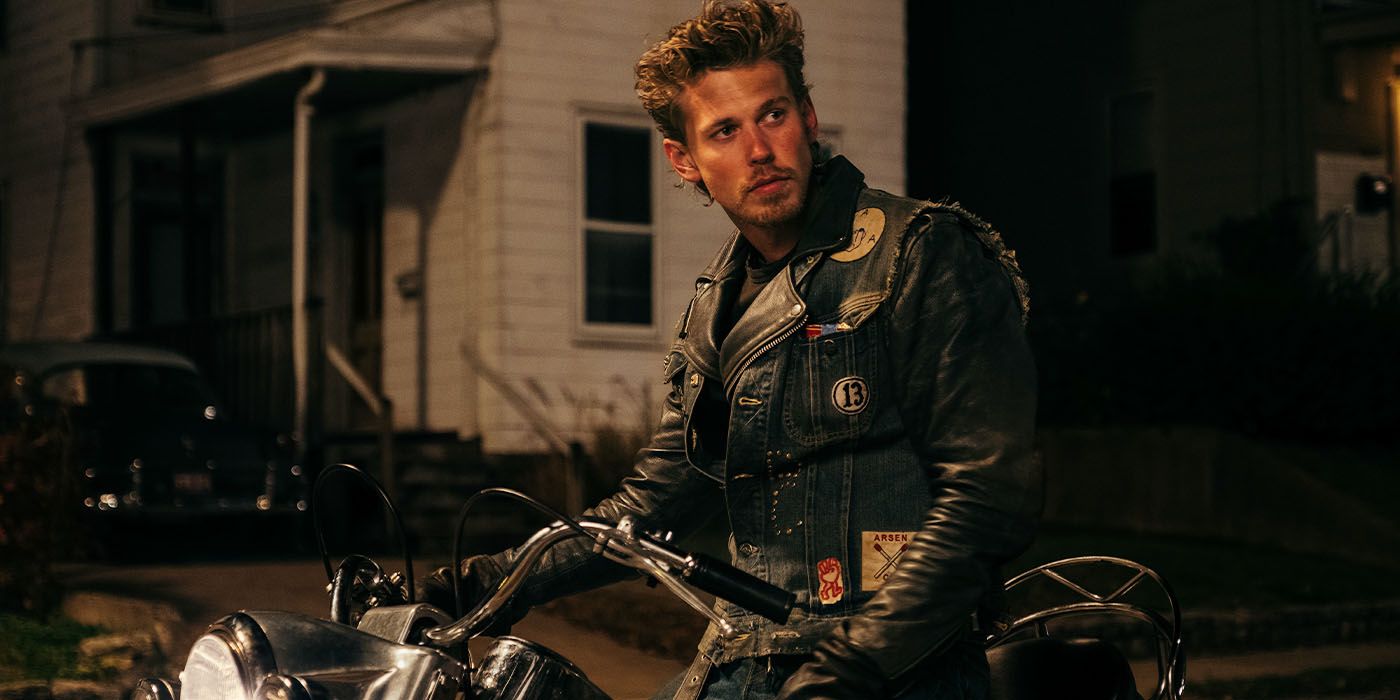 Austin Butler standing over a motorcycle looking off-camera in a denim vest and riding leathers