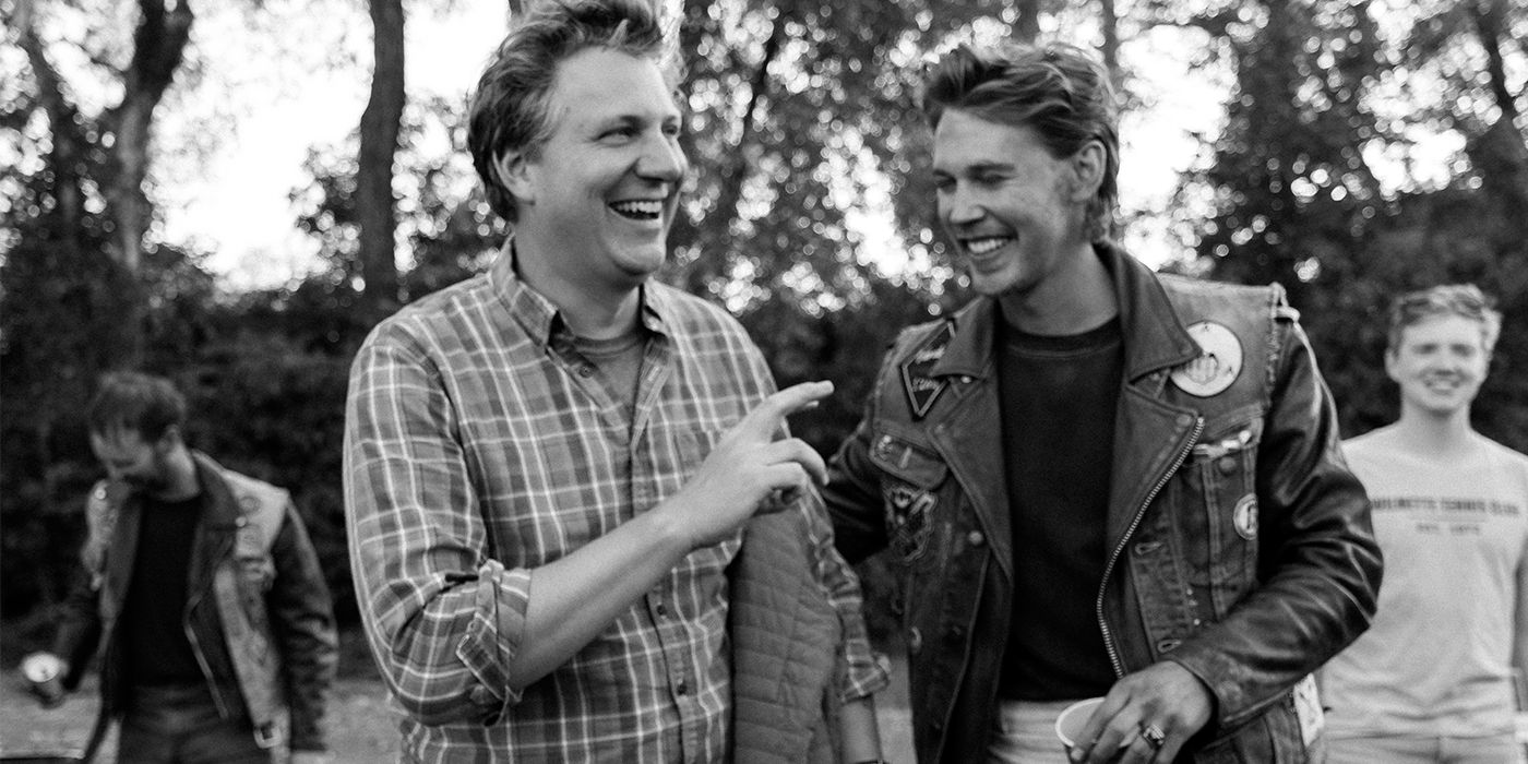 Black and white photo of Jeff Nichols and Austin Butler laughing on set in front of motorcycles and cast