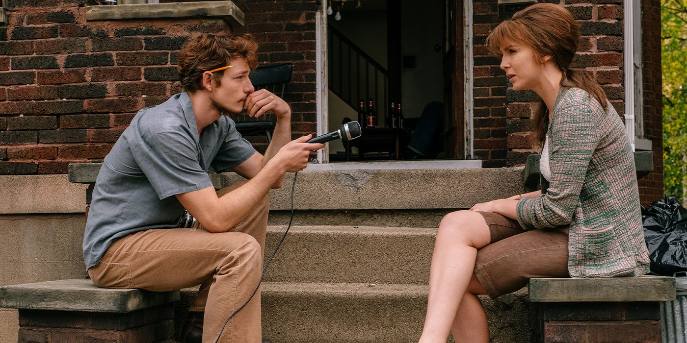 Mike Faist leans forward and points a microphone at Jodie Comer, who is sitting on a porch listening to her