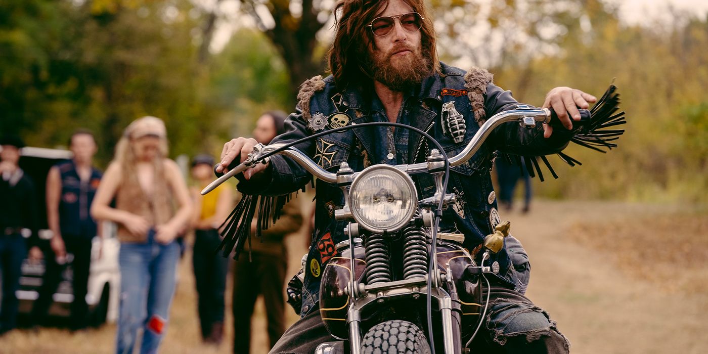 Norman Reedus sits on a motorcycle glaring off-camera in sunglasses with people in the background