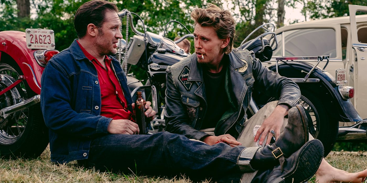 Tom Hardy and a barefoot Austin Butler sitting on the grass talking in front of motorcycles