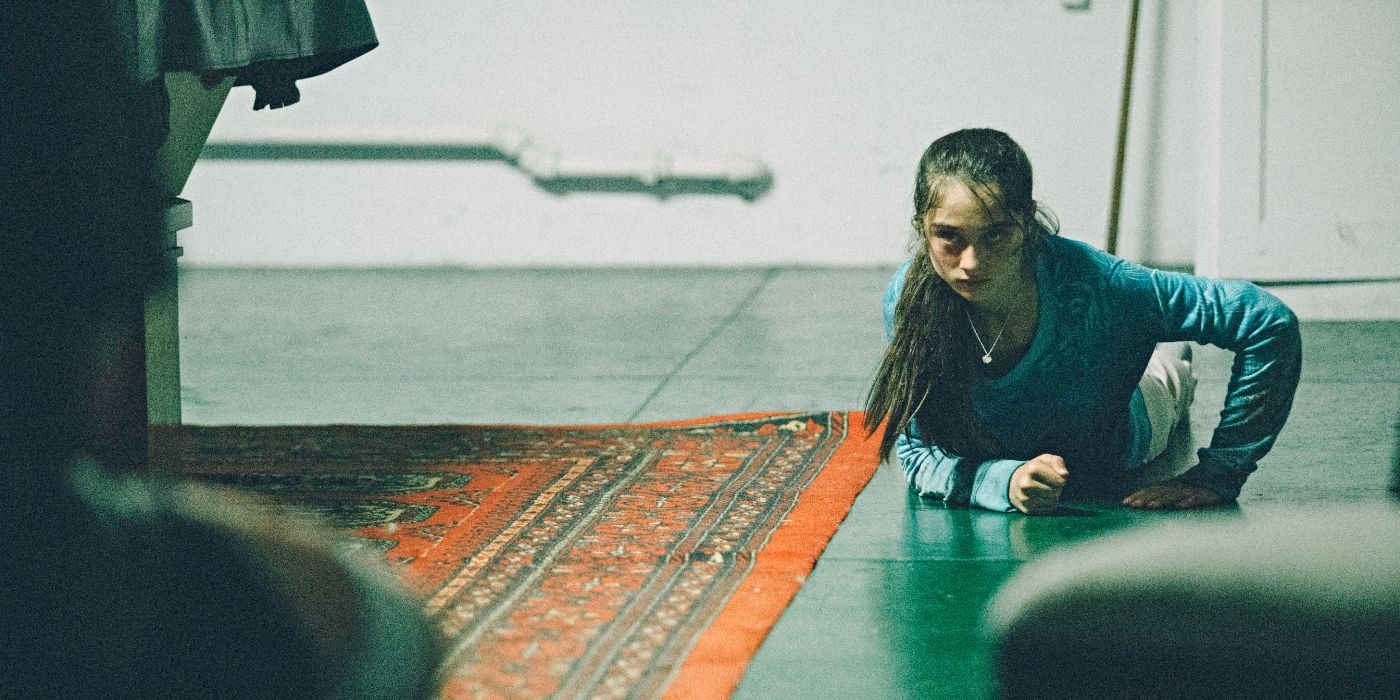 Raffey Cassidy as Kim Murphy, dragging herself across a tile floor next to a rug, looking serious in The Killing of a Sacred Deer