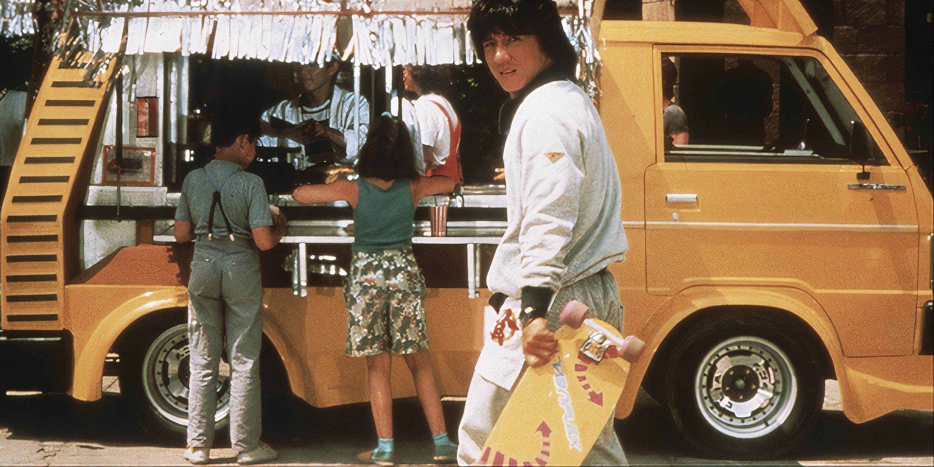 Jack Chan, holding a skateboard, looking back at the camera in Wheels on Meals.
