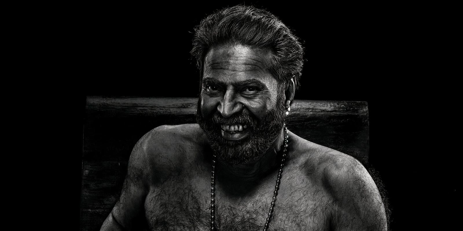 A hairy shirtless man in black and white wearing a necklace with a sinister toothy grin