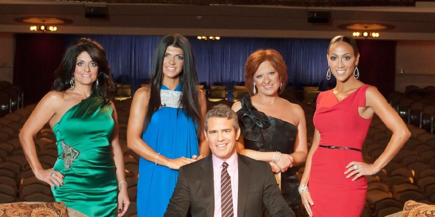 Group photo of The Real Housewives of New Jersey cast members: Kathy Wakile, Teresa Giudice, Caroline Manzo, and Melissa Gorga stand behind Andy Cohen, all dressed elegantly and smiling, in a theater setting.