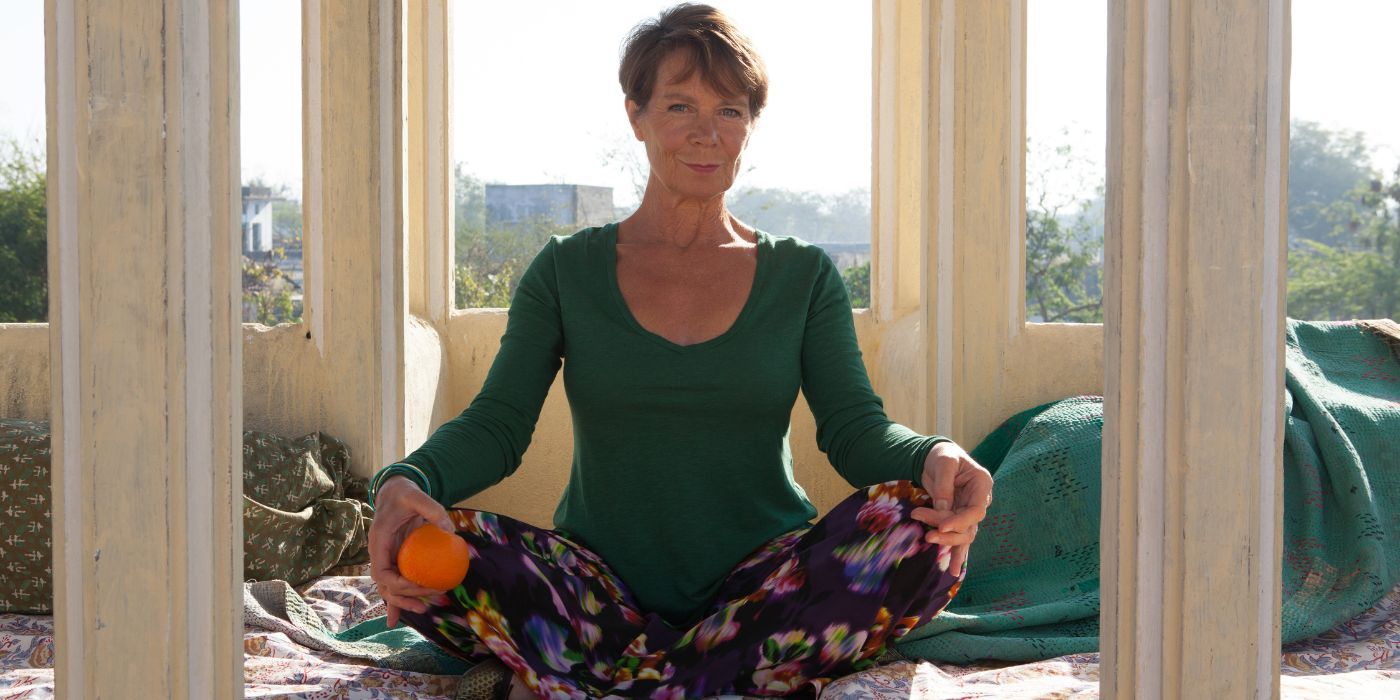 Celia Imrie sitting cross legged on a tarp holding an orange as Madge Hardcastle