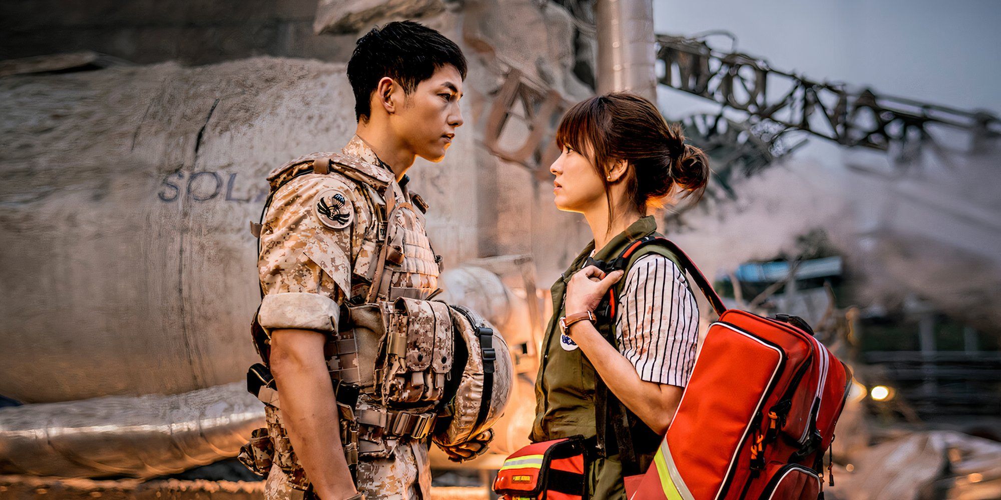 A couple look at each other in a war zone in Descendants of the Sun.