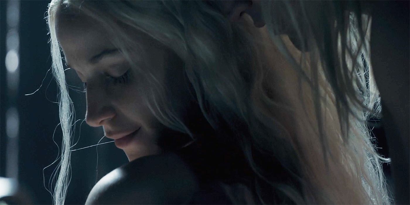 A blonde woman with no clothes on whom we can only see from the shoulders up is facing away from us but looking to the left. A man that we can hardly see approaches her from the back.