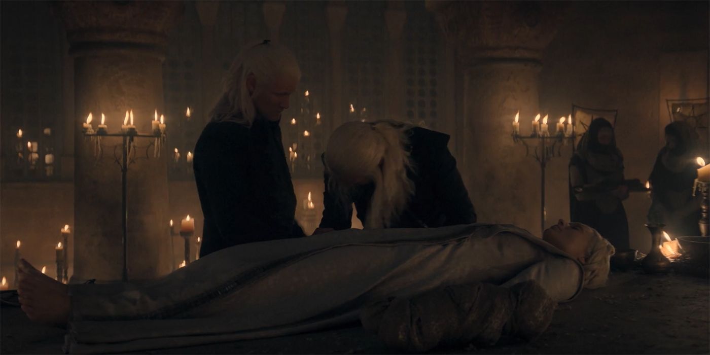 In a candle-lit room, an older blonde man (Viserys) leans in sorrow over a blonde woman (Aemma) wearing a grey dress. A man (Daemon) with blonde hair stands to his left and looks down at him.