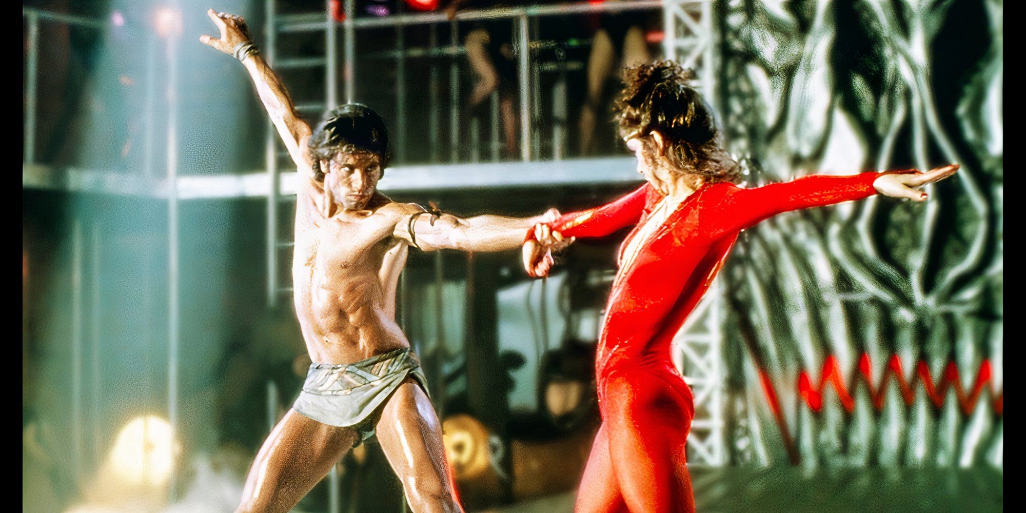 a shirtless man dancing with a woman in a red jumpsuit in Staying Alive