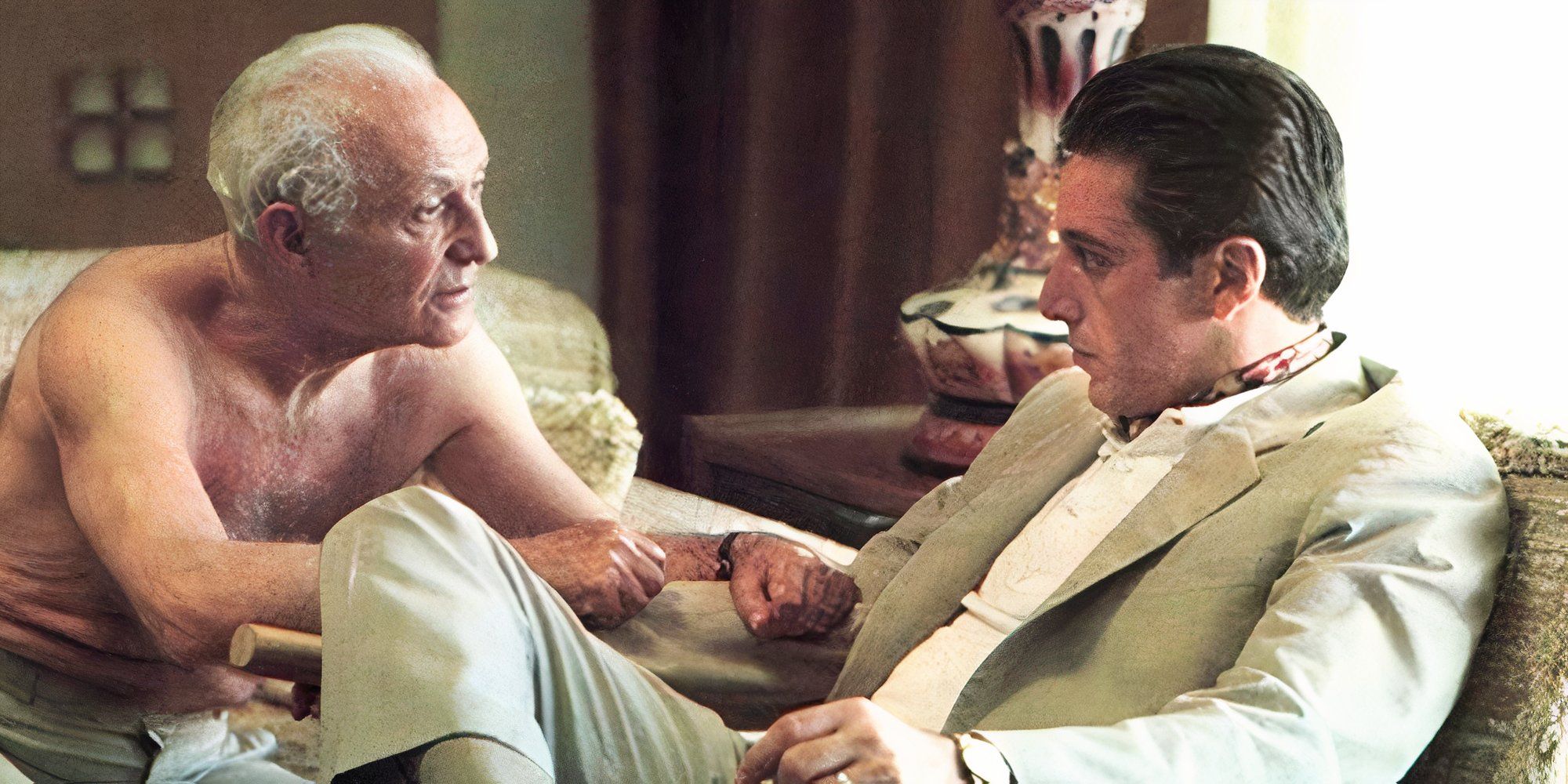 Lee Strasberg as Hyman Roth sitting on a couch leaning in towards Al Pacino as Michael Corleone who is sitting in a chair across from him in The Godfather: Part II.