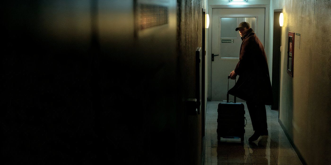 eddie redmayne wears a baseball hat and a long jacket while wheeling his suitcase down the hall