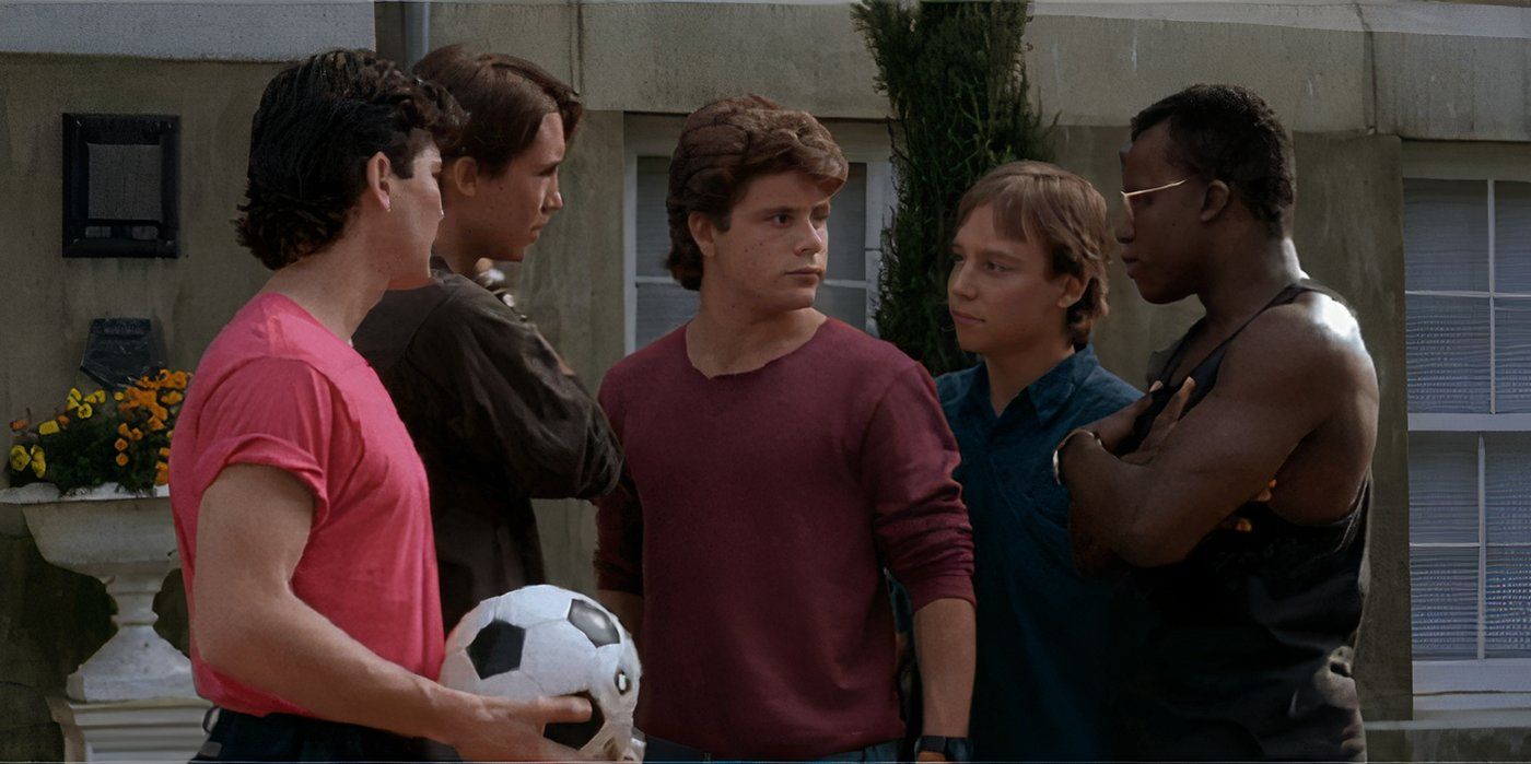 Five school-age young men stand in a cluster with arms folded, one holds a soccer ball