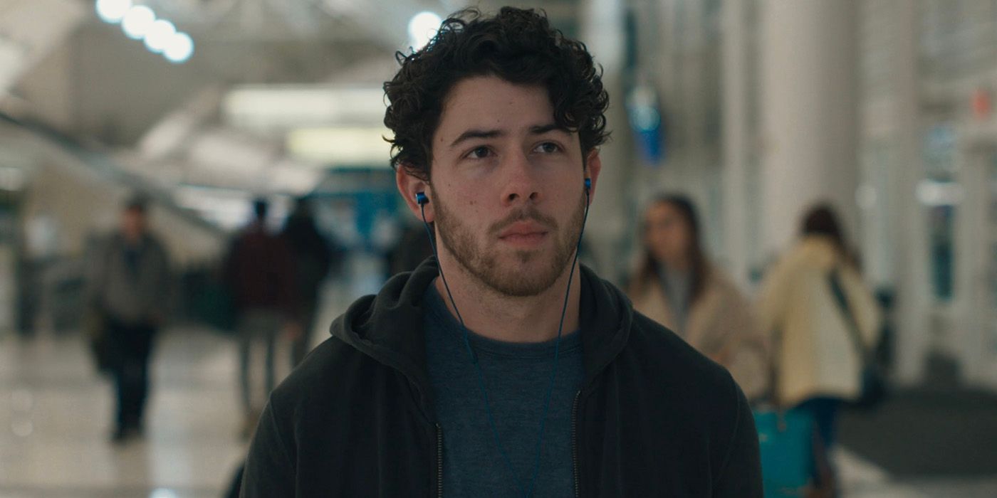 Nick Jonas walking through an airport with wired headphones on in The Good Half