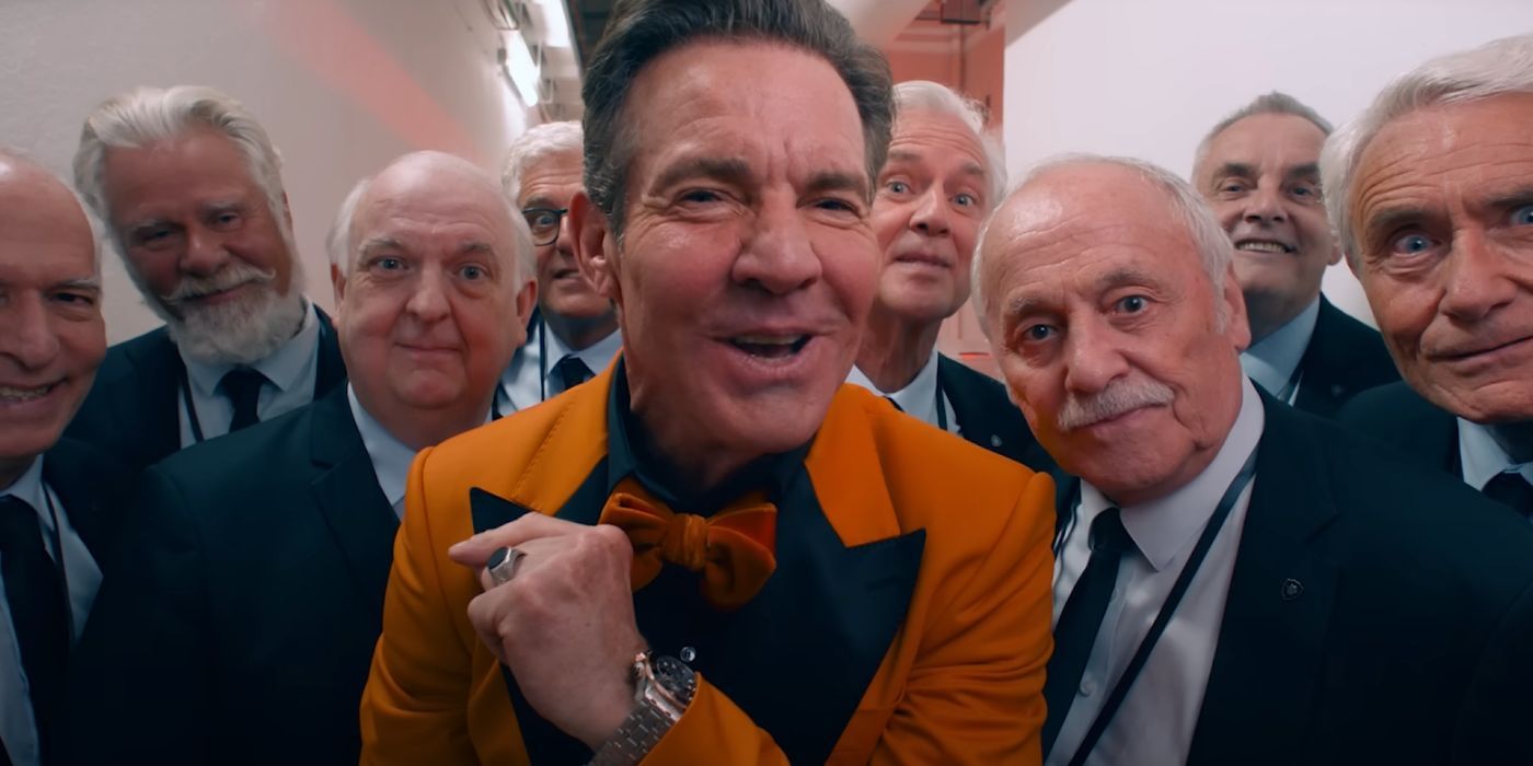 Dennis Quaid wearing an orange suit and making a unhinged smile while being surrounded by old white men in The Substances.