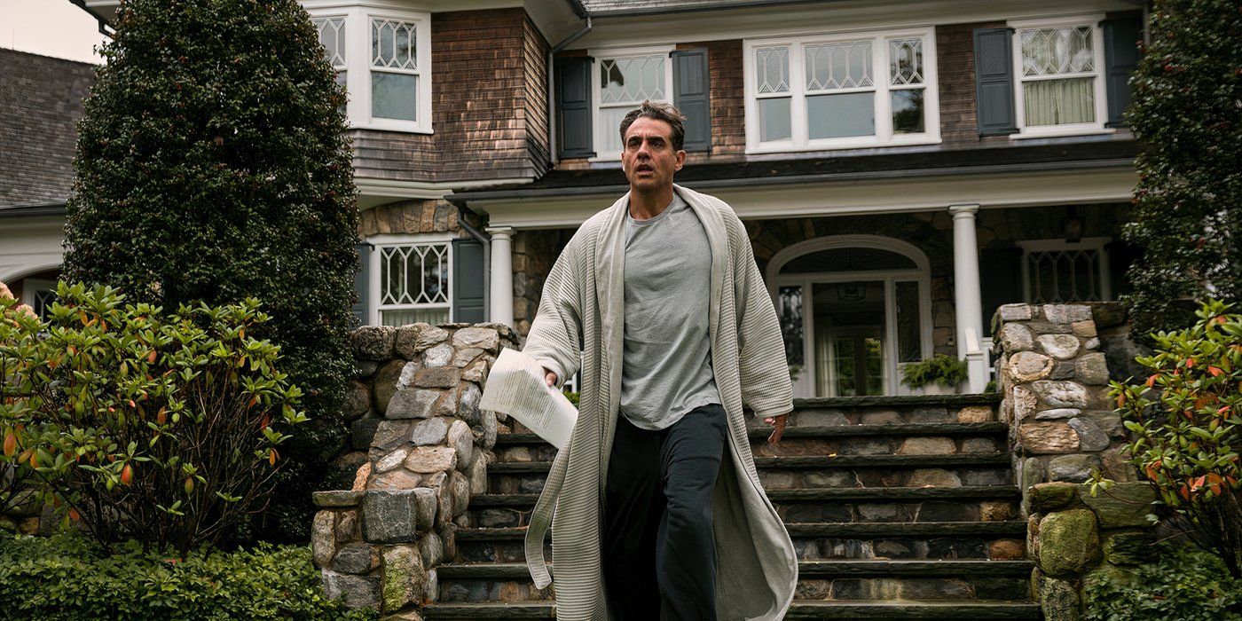 A man stands in front of a mansion in a robe holding a better and looking scared in The Watcher.