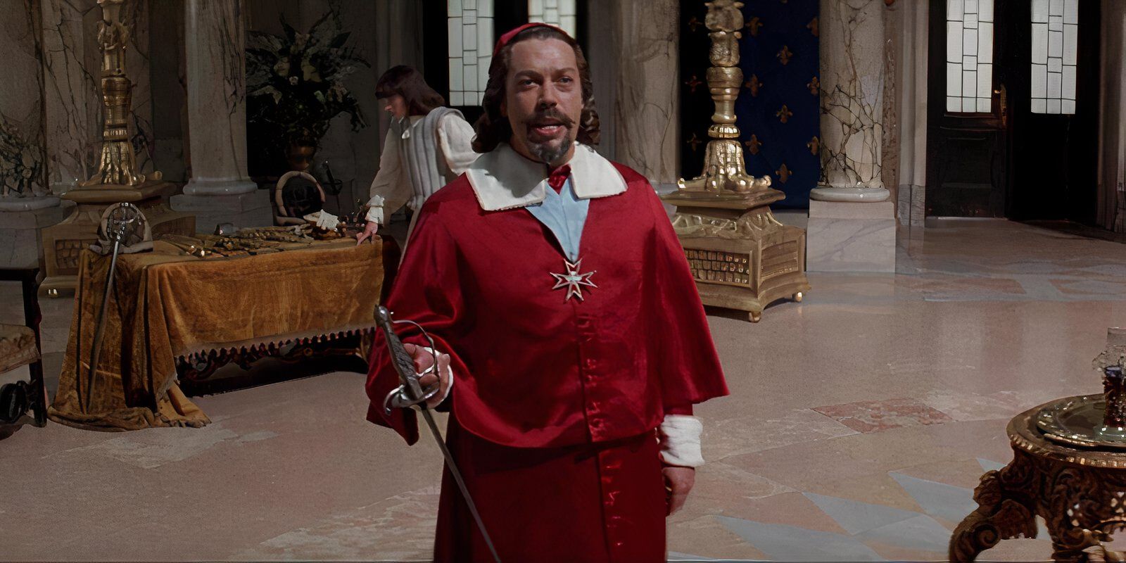 Tim Curry as Cardinal Richelieu wearing a bright red Cardinal outfit and holding a rapier sword while standing in a church in 'The Three Musketeers' (1993)