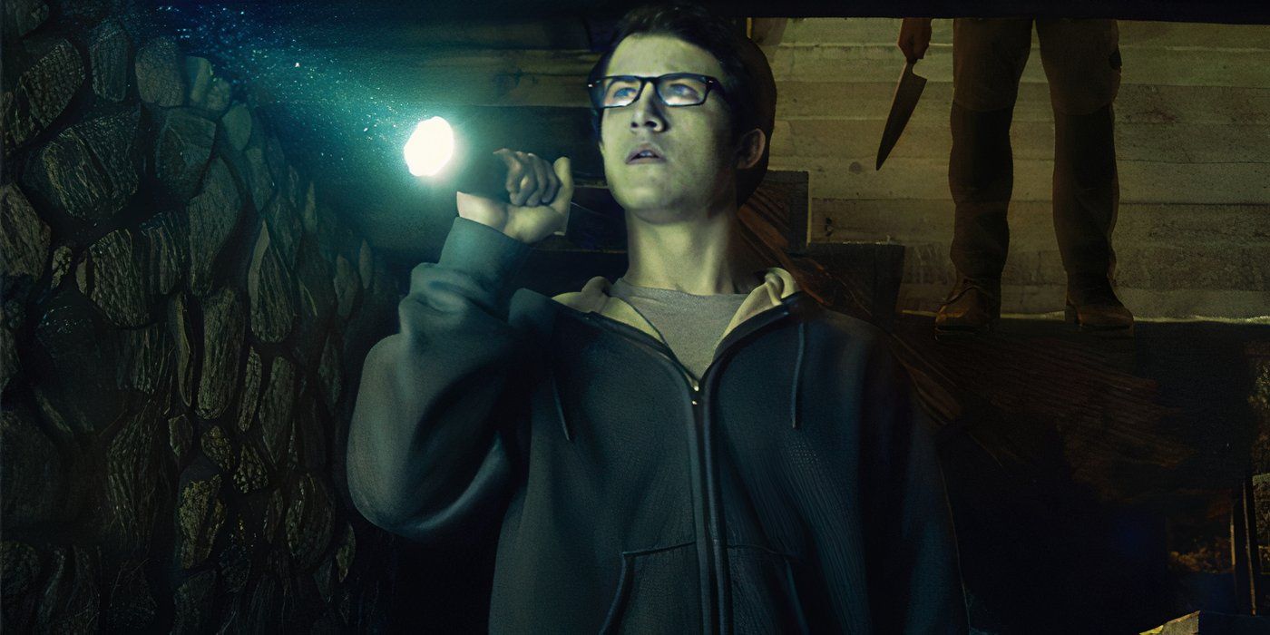 Dylan Minnette as Logan shines a flashlight in a dark room as a shadowy, knife-wielding figure appears behind him