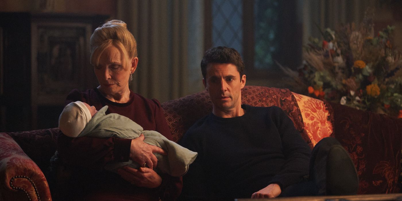 Ysabeau de Clermont (Lindsay Duncan) sitting on a couch and holding a newborn baby with Matthew Clairmont (Matthew Goode) sitting on her right in A Discovery of Witches