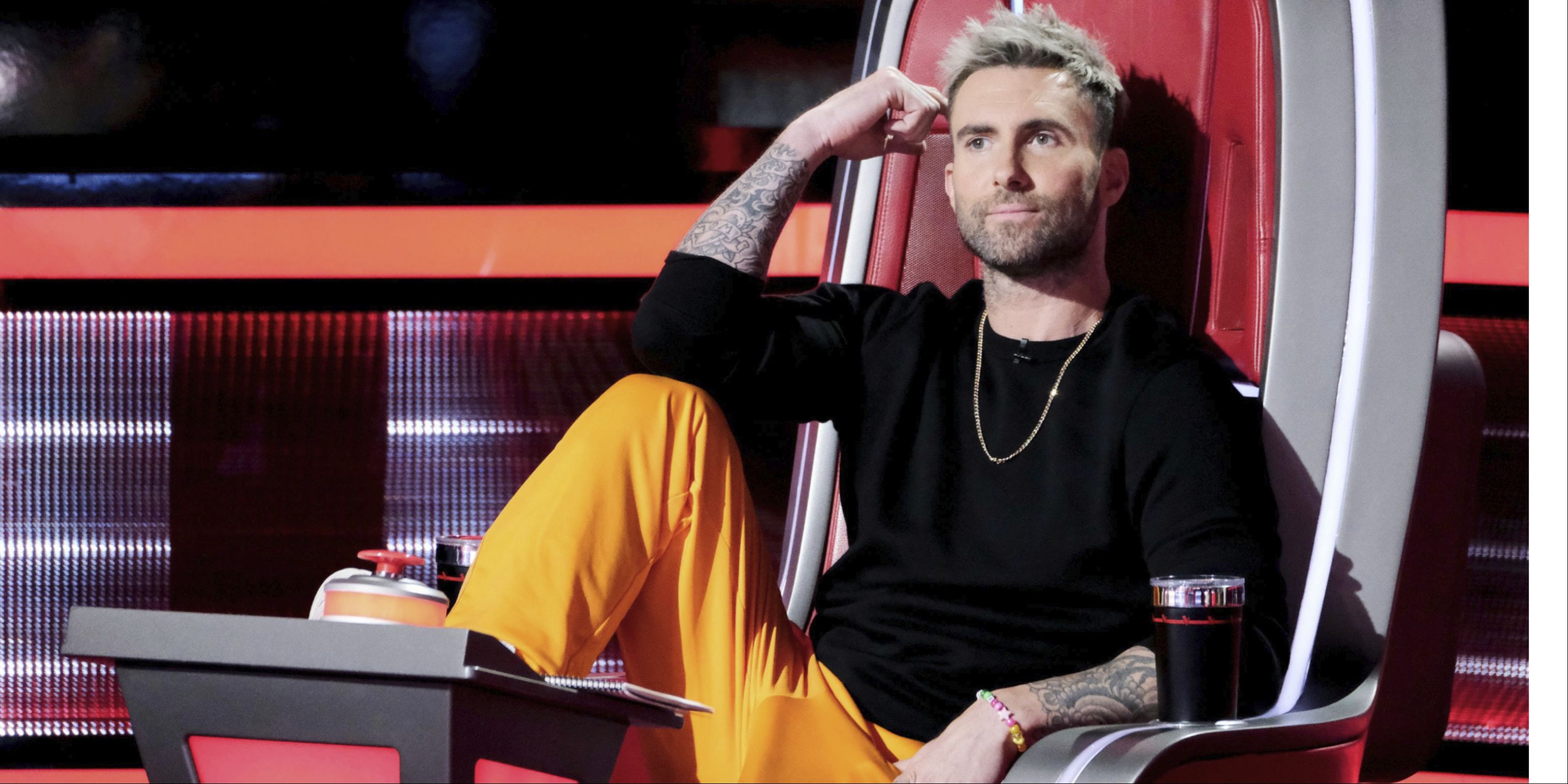 Adam Levine sitting in his chair on The Voice, leg up and arm to his head smiling.