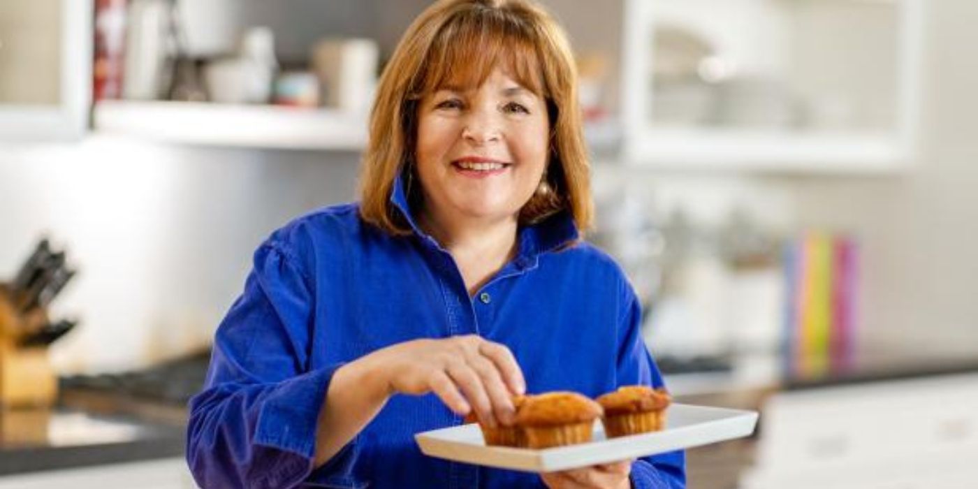 Ina Garten wears a bright blue shirt and stands in a kitchen holding a small plate of muffins in Barefoot Contessa.